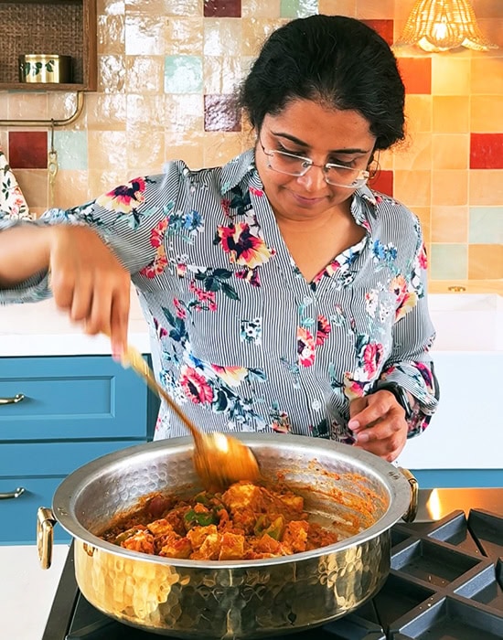 a woman stirring vegetables with paneer in a pan standing in her kitchen