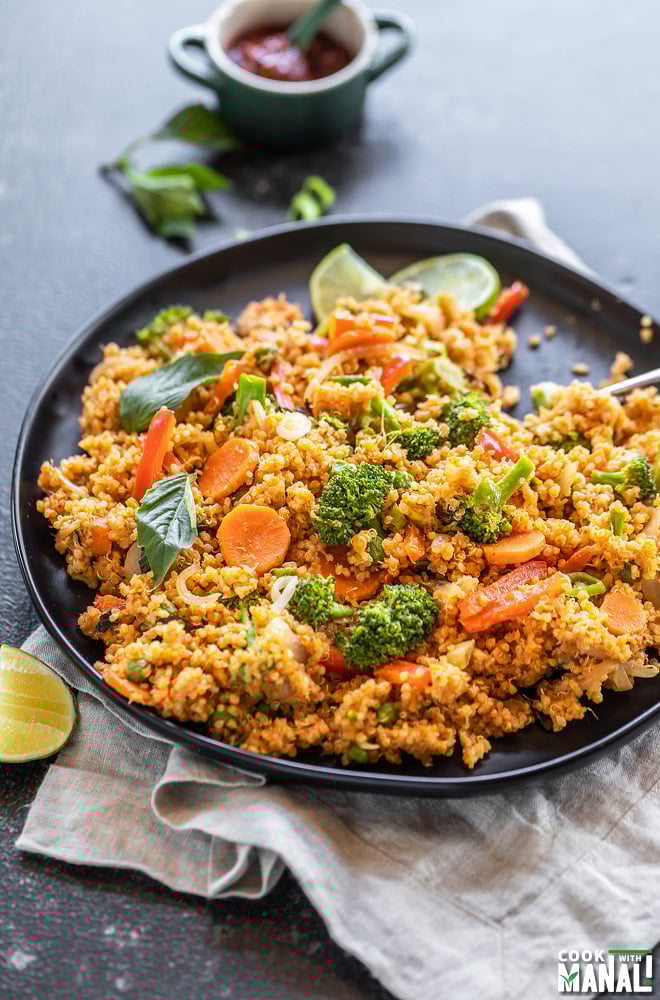 black plate with quinoa, veggies, basil, lime wedge and more basil and a bowl of hot sauce in the background
